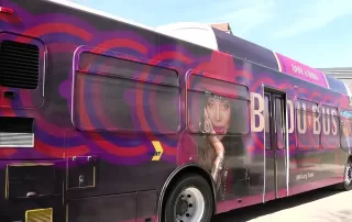 A large bus wrapped in vibrant purple and red circular patterns featuring a large image of Erykah Badu, a well-known singer, on the side. The bus is branded as the 'Badu Bus' with the words 'DART X BADU' on the top,in a collaboration with Dallas Area Rapid Transit (DART). The bus stands in an outdoor setting under a clear sky.