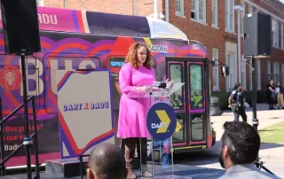 A large bus wrapped in vibrant purple and red circular patterns featuring a large image of Erykah Badu, a well-known singer, on the side. The bus is branded as the 'Badu Bus' with the words 'DART X BADU' on the top,in a collaboration with Dallas Area Rapid Transit (DART). The bus stands in an outdoor setting under a clear sky.