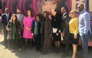 A large bus wrapped in vibrant purple and red circular patterns featuring a large image of Erykah Badu, a well-known singer, on the side. The bus is branded as the 'Badu Bus' with the words 'DART X BADU' on the top,in a collaboration with Dallas Area Rapid Transit (DART). The bus stands in an outdoor setting under a clear sky.