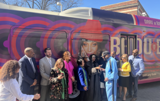 A large bus wrapped in vibrant purple and red circular patterns featuring a large image of Erykah Badu, a well-known singer, on the side. The bus is branded as the 'Badu Bus' with the words 'DART X BADU' on the top,in a collaboration with Dallas Area Rapid Transit (DART). The bus stands in an outdoor setting under a clear sky.