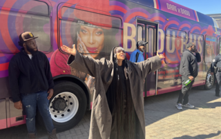 A large bus wrapped in vibrant purple and red circular patterns featuring a large image of Erykah Badu, a well-known singer, on the side. The bus is branded as the 'Badu Bus' with the words 'DART X BADU' on the top,in a collaboration with Dallas Area Rapid Transit (DART). The bus stands in an outdoor setting under a clear sky.