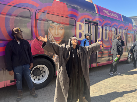 A large bus wrapped in vibrant purple and red circular patterns featuring a large image of Erykah Badu, a well-known singer, on the side. The bus is branded as the 'Badu Bus' with the words 'DART X BADU' on the top,in a collaboration with Dallas Area Rapid Transit (DART). The bus stands in an outdoor setting under a clear sky.