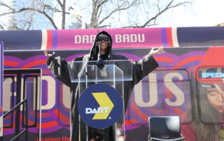 A large bus wrapped in vibrant purple and red circular patterns featuring a large image of Erykah Badu, a well-known singer, on the side. The bus is branded as the 'Badu Bus' with the words 'DART X BADU' on the top,in a collaboration with Dallas Area Rapid Transit (DART). The bus stands in an outdoor setting under a clear sky.