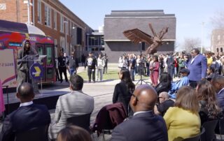 A large bus wrapped in vibrant purple and red circular patterns featuring a large image of Erykah Badu, a well-known singer, on the side. The bus is branded as the 'Badu Bus' with the words 'DART X BADU' on the top,in a collaboration with Dallas Area Rapid Transit (DART). The bus stands in an outdoor setting under a clear sky.